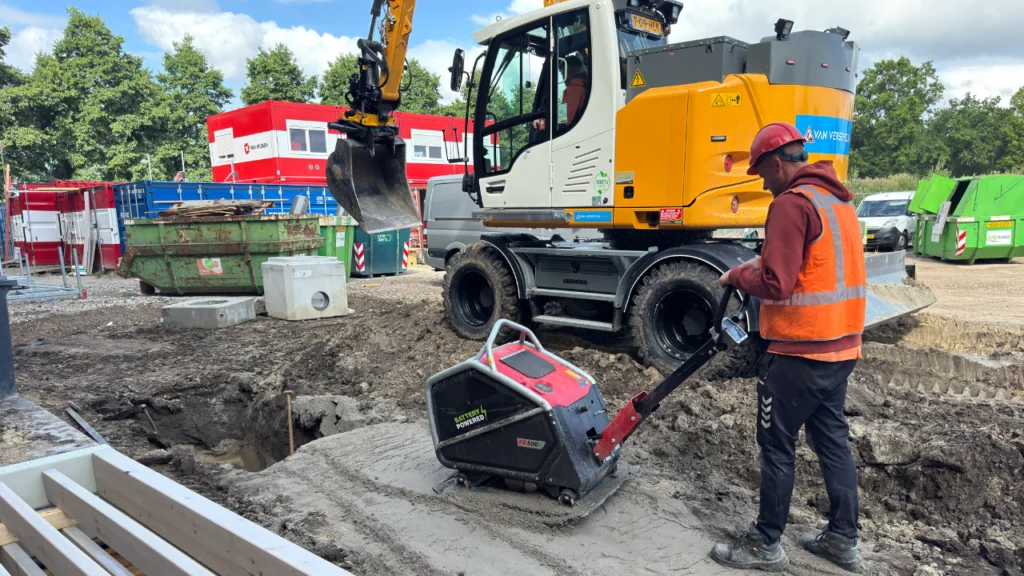 Medewerker met een zware Swepac accu-trilplaat naast een graafmachine