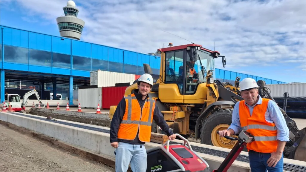 Twee medewerkers met een trilplaat op een werkterrein bij een luchthaven