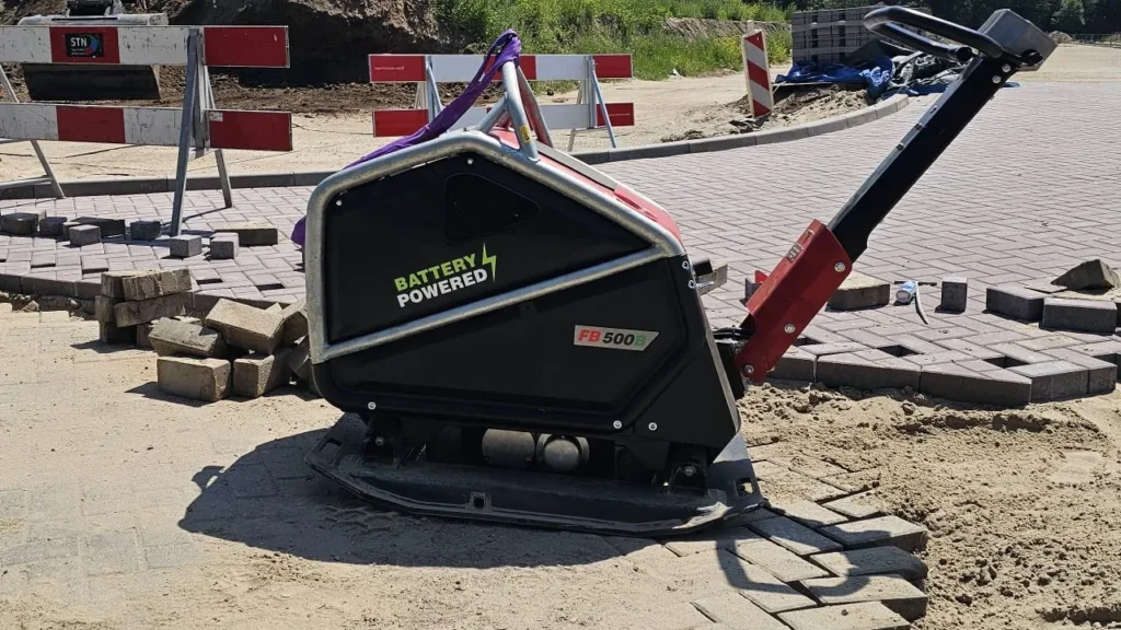 stratenmaker gebruikt een accu trilplaat in een smalle woonstraat tussen geparkeerde auto’s en rijtjeshuizen