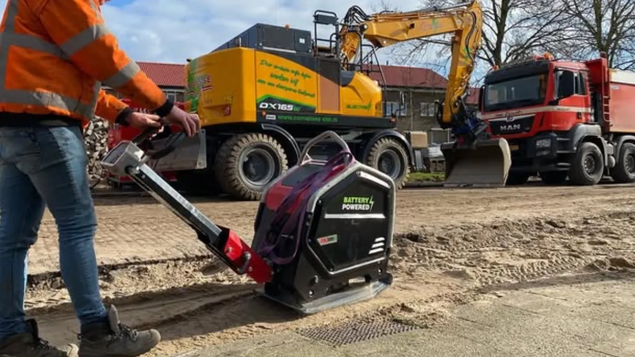 bouwvakker bedient een accu-trilplaat op een stedelijke bouwplaats met mobiele laadunit op de achtergrond